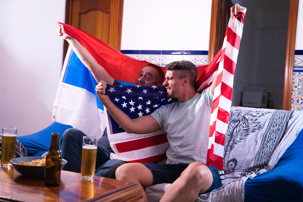 Два друга-мужчины и любители спорта смотрят спортивное событие по телевизору, выпивая пиво. Каждый из мужчин несет флаг на своих плечах. Спортивная концепция - Фото, изображение