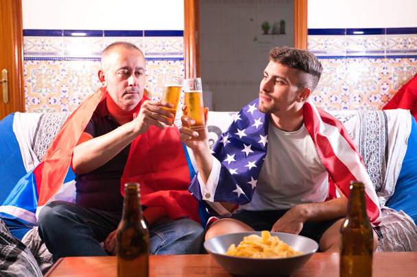 Twee mannelijke vrienden en sportfans kijken een sportevenement op televisie terwijl ze toosten met een biertje. Elk van de mannen draagt een vlag op zijn schouders. Sportconcept - Foto, afbeelding