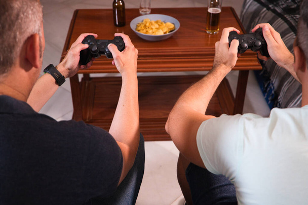 ビデオコンソールのコントローラーを持っている2人の友人の手の詳細は、男性は軽食とビールを飲みながらビデオゲームをしています。ビデオゲームと友情の概念. - 写真・画像