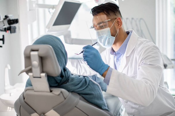 医療用マスクおよびフェイスシールドにおける中東男性歯科医患者の歯をチェック - 写真・画像
