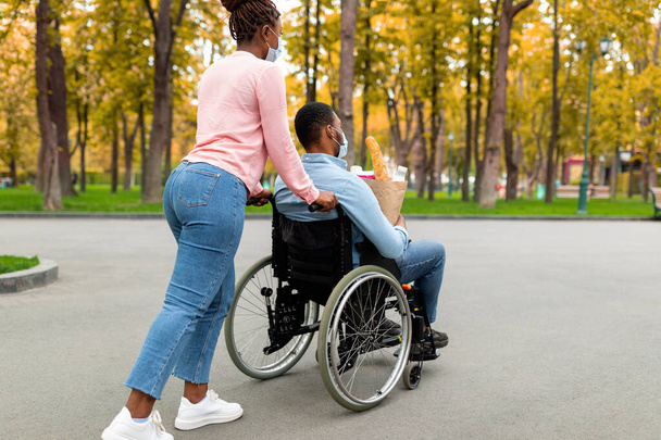 Mladý černošky zdravotně postižený chlap na vozíku drží papírový sáček s jídlem, vracející se domů z nákupů s manželkou během covid - Fotografie, Obrázek