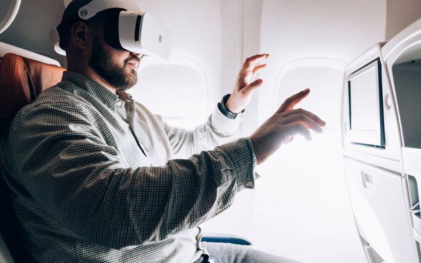 Man in VR glasses projecting 3d dimension during aircraft flight in business class with wifi connection, millennial male passenger testing imaginary environment while watching 3d video cinema - Photo, Image