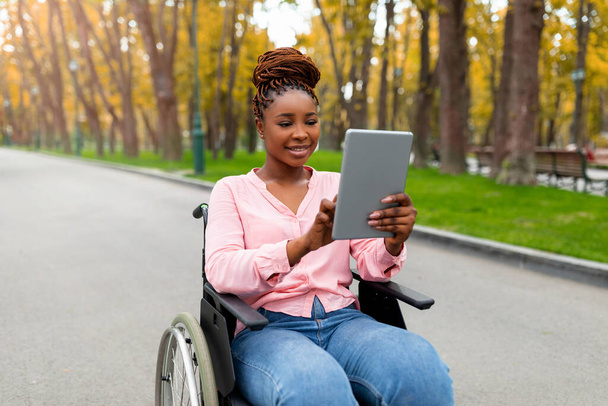 秋の公園で遠隔コミュニケーションのためにタブレットPCを使用して車いすの中で障害のある若い黒人女性の肖像画 - 写真・画像