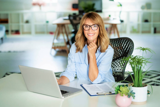 Sorrindo empresária de meia idade trabalhando laptop enquanto sentado na mesa no escritório.  - Foto, Imagem
