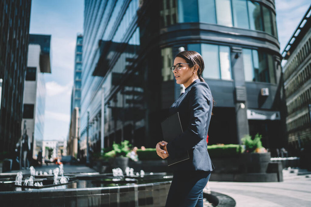 Side view of serious young woman in formal suit and glasses with paperwork waiting for meeting with partner standing in street of modern city looking away - Photo, image