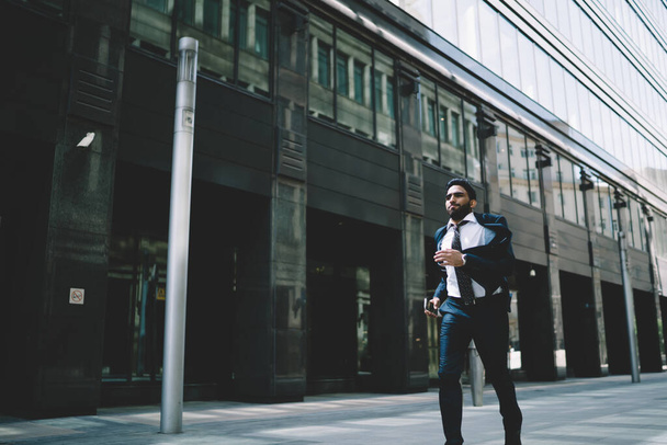 etniczny brodaty wykonawczy mężczyzna z czarnymi włosami ubrany w formalny garnitur z krawatem chodzący szybko z telefonem komórkowym w ręku w mieście - Zdjęcie, obraz