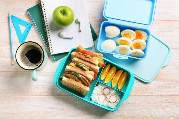 Lunch boxes with tasty food, cup of coffee, earphones and stationery on light wooden background - Photo, Image