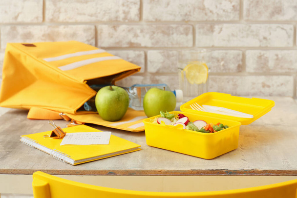 Lunch box with tasty vegetables, bag, apples and stationery on table against brick wall - Foto, immagini