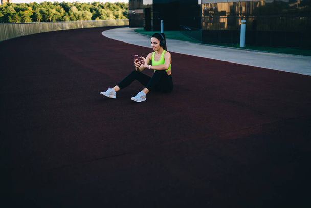  Young slim woman in active wear training on stadium doing squats exercises for lower body strength,caucasian girl keeping fitness goals having workout for energy wellness listen music in headphones - Foto, Imagen