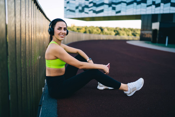  Young slim woman in active wear training on stadium doing squats exercises for lower body strength,caucasian girl keeping fitness goals having workout for energy wellness listen music in headphones - Photo, Image