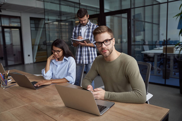 Młody biały mężczyzna, pracownik biurowy korzystający z laptopa, siedzący przy biurku i patrzący w kamerę. Grupa młodych wielokulturowych przedsiębiorców współpracujących w nowoczesnym biurze - Zdjęcie, obraz