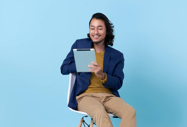 Businessman asian happy smiling using a digital tablet while sitting on chair isolated on bright blue background.	 - Zdjęcie, obraz