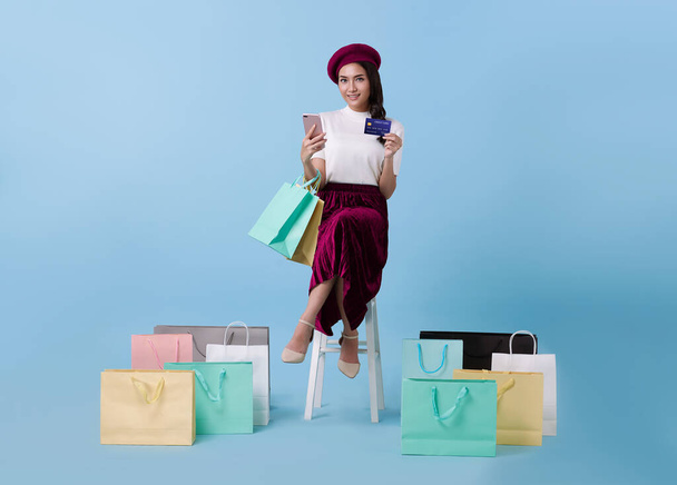 Güzel Asyalı kadın alışverişçi oturuyor ve elinde mavi arka planda kredi kartı ve cep telefonu ile alışveriş torbaları taşıyor.. - Fotoğraf, Görsel