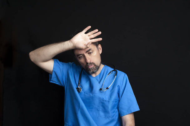 Médecin vêtu d'un uniforme bleu d'hôpital, sombre et barbu, portant un stéthoscope autour du cou, essuyant son front avec sa main avec une expression fatiguée ou souffrante.Un fond noir derrière lui - Photo, image