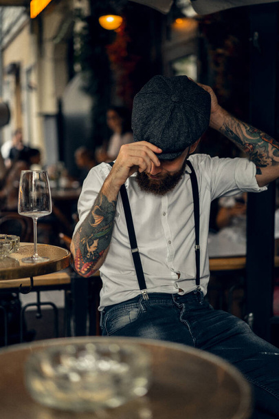 Fiatal szakállas férfi egy kávézóban az utcán egy pohár borral. Romantikus fickó fehér póló sapkában és nadrágtartóban a városban. Csúcsszeműek. régi fassion retro. - Fotó, kép