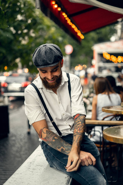 Young bearded man in a cafe on the street smokes a cigarette. Romantic guy in a white shirt cap and suspenders in the city. Peaky Blinders. old fashionable retro. - Foto, Imagen