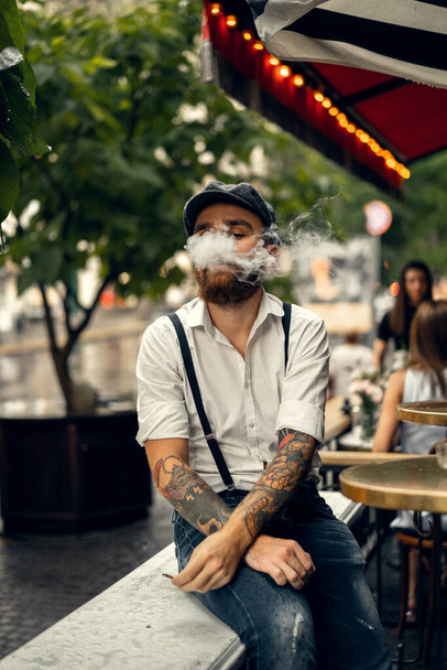 Fiatal szakállas férfi egy kávézóban az utcán cigarettázik. Romantikus fickó fehér póló sapkában és nadrágtartóban a városban. Csúcsszeműek. régimódi retro. - Fotó, kép