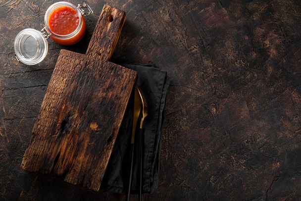  Old wooden cutting board, a jar of tomato sauce and cutlery on a dark background. Food background with free space for text - Photo, Image