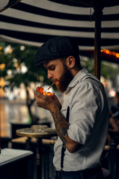 Young bearded man in a cafe on the street smokes a cigarette. Romantic guy in a white shirt cap and suspenders in the city. Peaky Blinders. old fashionable retro. - Foto, imagen