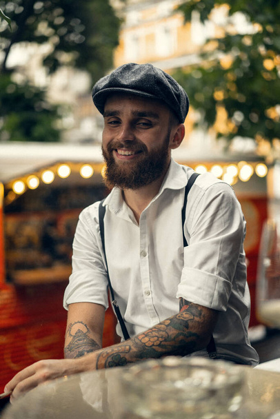 Young bearded man in a cafe on the street smokes a cigarette. Romantic guy in a white shirt cap and suspenders in the city. Peaky Blinders. old fashionable retro. - Foto, imagen