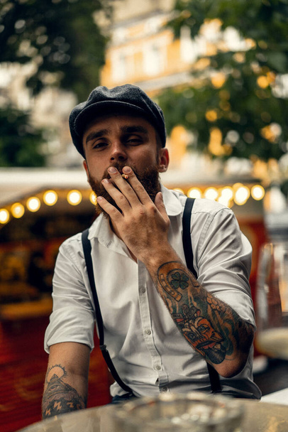 Young bearded man in a cafe on the street smokes a cigarette. Romantic guy in a white shirt cap and suspenders in the city. Peaky Blinders. old fashionable retro. - Fotó, kép