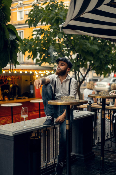Young bearded man in a cafe on the street with a glass of wine. Romantic guy in a white shirt cap and suspenders in the city. Peaky Blinders. old fassion retro. - Valokuva, kuva