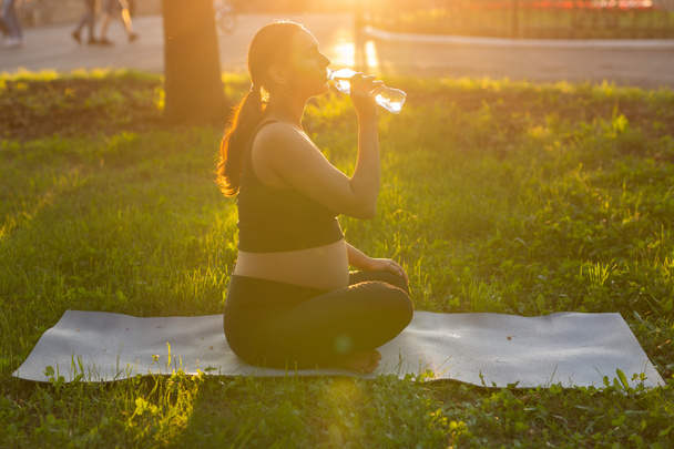 妊娠,スポーツと健康のライフスタイル – ヨガの後の晴れた夏の日に芝生の上に座ってボトルから水を飲む妊婦 - 写真・画像