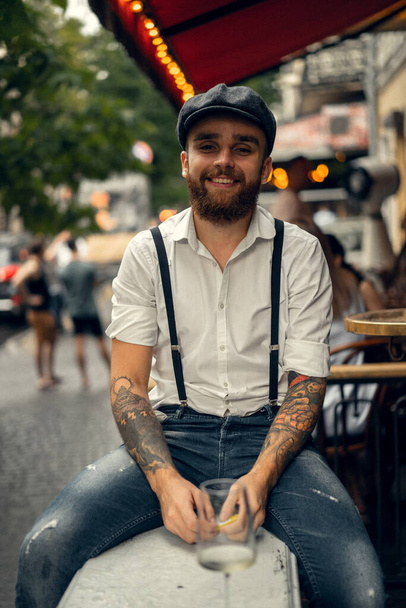 Young bearded man in a cafe on the street with a glass of wine. Romantic guy in a white shirt cap and suspenders in the city. Peaky Blinders. old fassion retro. - 写真・画像