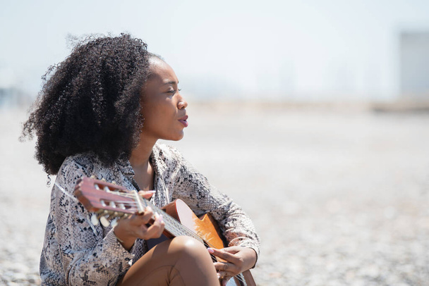 Relaxed young woman sitting on a gravel stone ground in the oper air playing her acoustic guitar and singing - Foto, Imagen