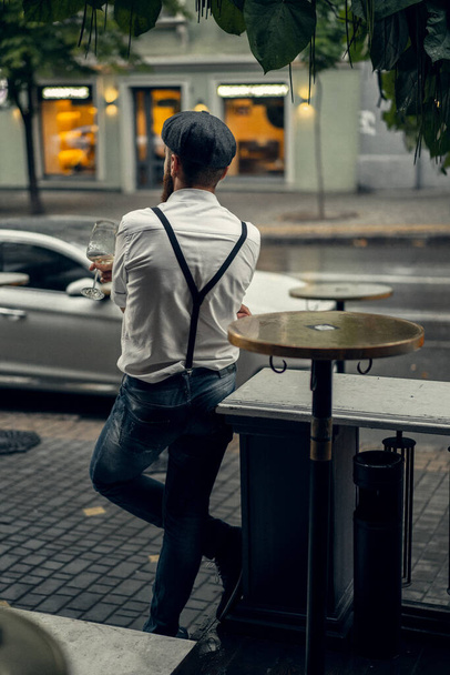 Young bearded man in a cafe on the street with a glass of wine. Romantic guy in a white shirt cap and suspenders in the city. Peaky Blinders. old fassion retro. - Photo, Image