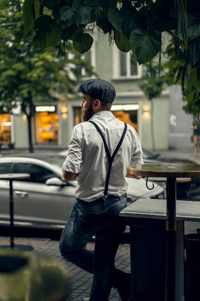 Young bearded man in a cafe on the street with a glass of wine. Romantic guy in a white shirt cap and suspenders in the city. Peaky Blinders. old fassion retro. - Photo, Image