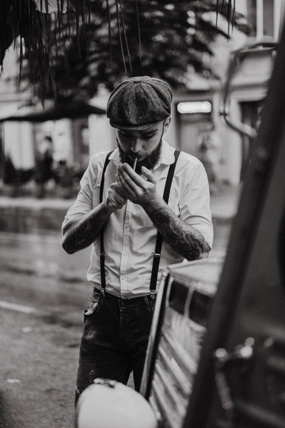 Young bearded tattoord man in a cafe on the street smokes a cigarette. Romantic guy in a white shirt cap and suspenders in the city. Peaky Blinders. old fashionable retro. - Photo, Image