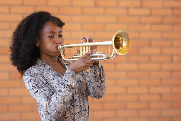 Retrato de una joven afroamericana tocando la trompeta afuera sobre un fondo de pared de ladrillo - Foto, imagen