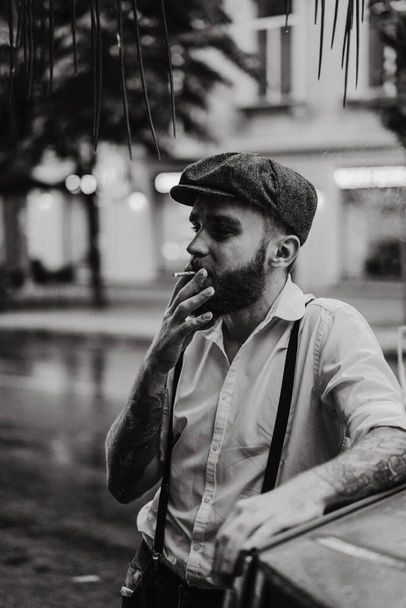 Young bearded tattoord man in a cafe on the street smokes a cigarette. Romantic guy in a white shirt cap and suspenders in the city. Peaky Blinders. old fashionable retro. - Foto, Bild