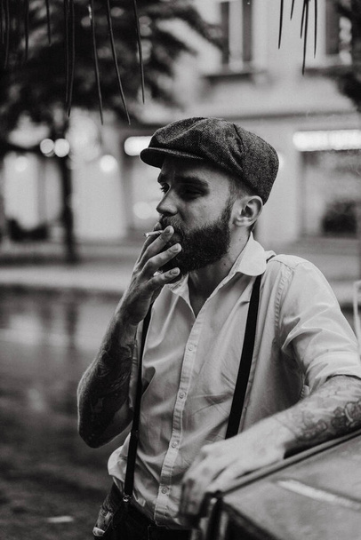 Young bearded tattoord man in a cafe on the street smokes a cigarette. Romantic guy in a white shirt cap and suspenders in the city. Peaky Blinders. old fashionable retro. - Foto, immagini
