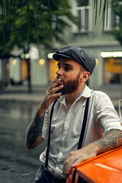 Young bearded tattoord man in a cafe on the street smokes a cigarette. Romantic guy in a white shirt cap and suspenders in the city. Peaky Blinders. old fashionable retro. - Fotografie, Obrázek