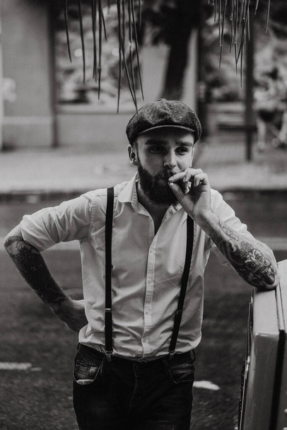 Young bearded tattoord man in a cafe on the street smokes a cigarette. Romantic guy in a white shirt cap and suspenders in the city. Peaky Blinders. old fashionable retro. - Photo, image