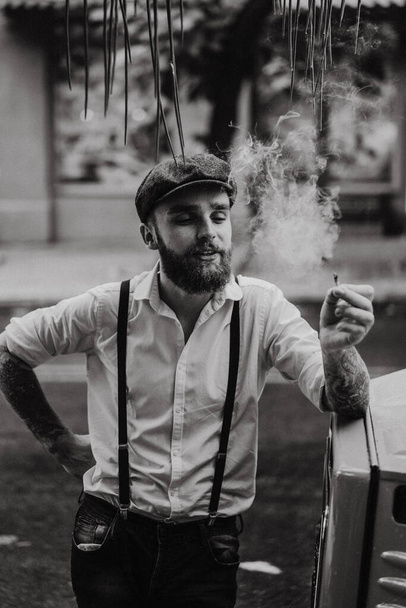 Young bearded tattoord man in a cafe on the street smokes a cigarette. Romantic guy in a white shirt cap and suspenders in the city. Peaky Blinders. old fashionable retro. - Foto, afbeelding