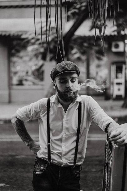 Young bearded tattoord man in a cafe on the street smokes a cigarette. Romantic guy in a white shirt cap and suspenders in the city. Peaky Blinders. old fashionable retro. - Zdjęcie, obraz