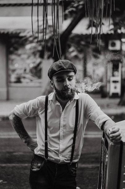 Young bearded tattoord man in a cafe on the street smokes a cigarette. Romantic guy in a white shirt cap and suspenders in the city. Peaky Blinders. old fashionable retro. - Foto, immagini