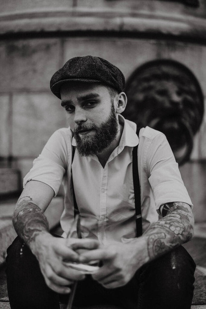 Young bearded tattooed man drinking wine from a glass on the street. Romantic guy in a white shirt, cap and suspenders in the city. Peaky Blinders. old-fashioned, retro. - Foto, immagini
