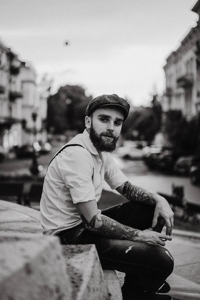 Young bearded tattooed man drinking wine from a glass on the street. Romantic guy in a white shirt, cap and suspenders in the city. Peaky Blinders. old-fashioned, retro. - Foto, Imagem