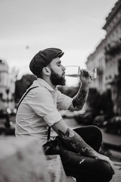 Young bearded tattooed man drinking wine from a glass on the street. Romantic guy in a white shirt, cap and suspenders in the city. Peaky Blinders. old-fashioned, retro. - Фото, изображение