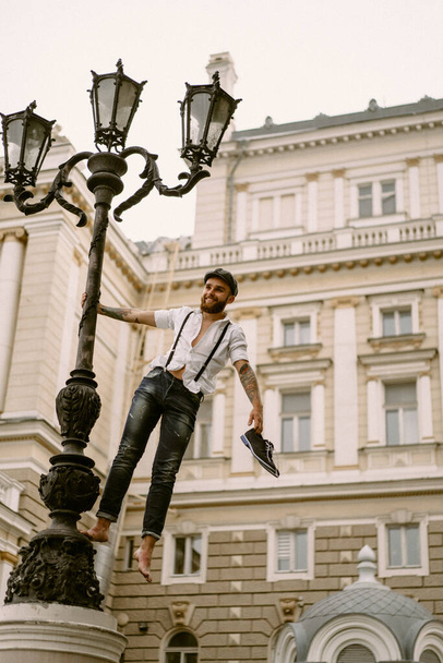 Young bearded tattooed man. A romantic guy in a white shirt, cap and suspenders walks in the city. revolves around a lamppost. Peaky Blinders. old-fashioned, retro. - Foto, Imagen