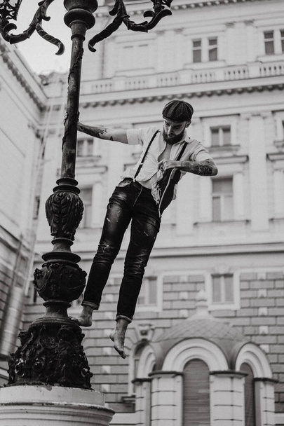 Young bearded tattooed man. A romantic guy in a white shirt, cap and suspenders walks in the city. revolves around a lamppost. Peaky Blinders. old-fashioned, retro. - Fotografie, Obrázek