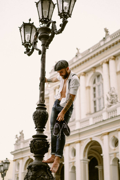 Young bearded tattooed man. A romantic guy in a white shirt, cap and suspenders walks in the city. revolves around a lamppost. Peaky Blinders. old-fashioned, retro. - Fotoğraf, Görsel