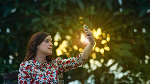 Beautiful young latin woman in floral design dress taking a selfie with her smartphone in the garden at sunset with the sun's rays passing through the trees in the background - Фото, зображення
