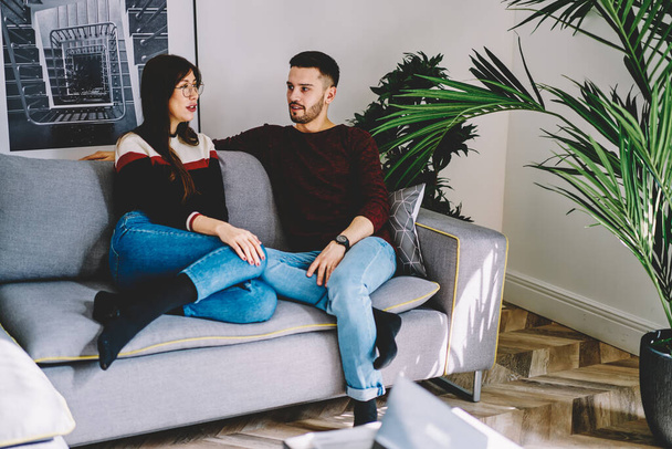 Young male and female friend sitting on sofa together communicating about hobby and leisure in living room, caucasian couple in love enjoying free time sitting at home interior talking to each other - Φωτογραφία, εικόνα