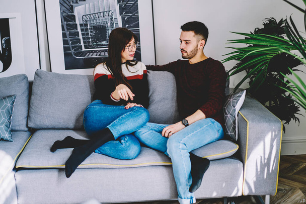 Positive male and female marriage sitting on sofa together communicating and resting on free time together, young marriage discussing relationships enjoying conversation in living room in apartment - Фото, изображение