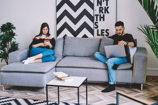 Millennial woman and man hipster friends concentrated on using digital devices on free time addicted to online chatting, young marriage spending free time at living room on gadgets ignoring each other - 写真・画像